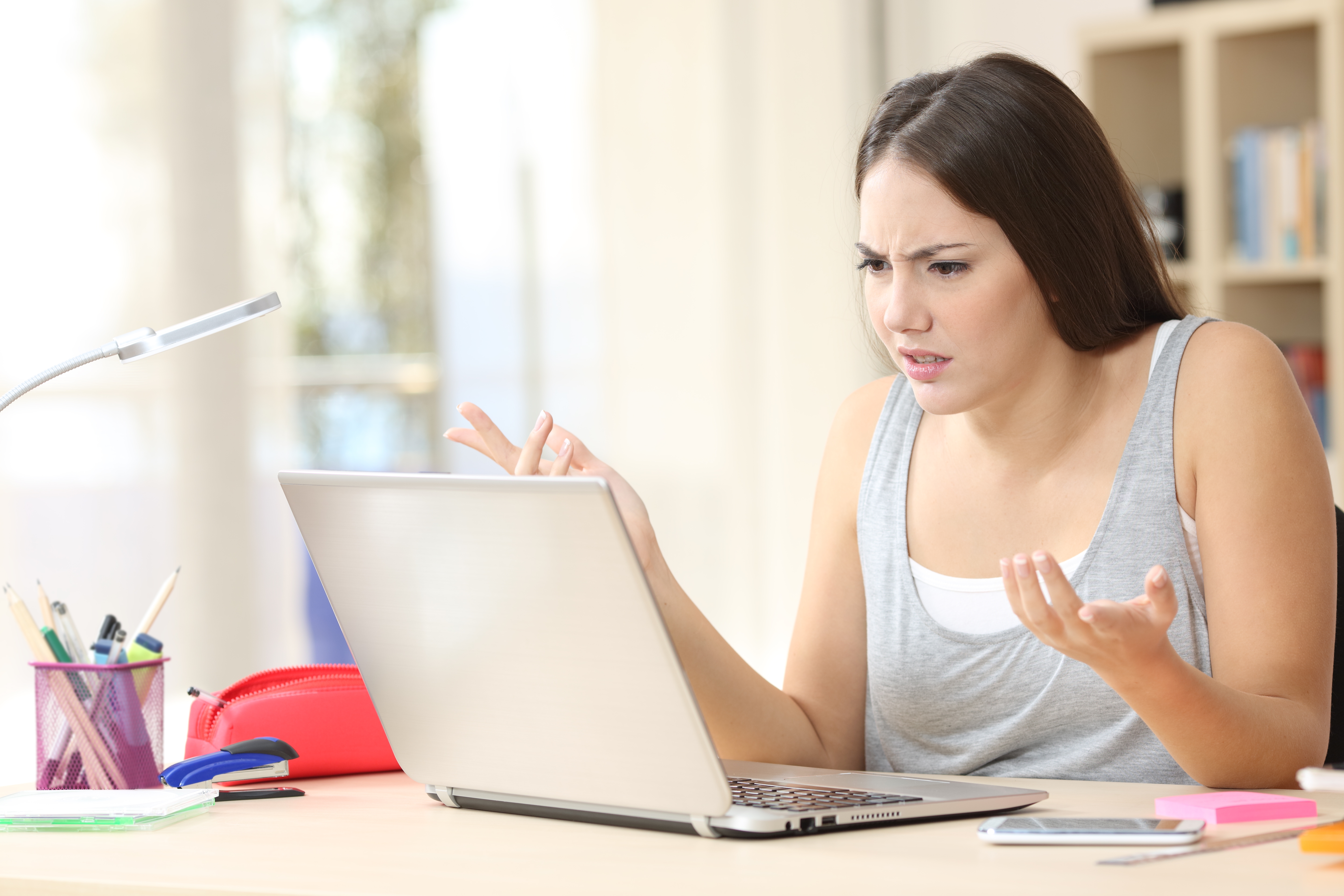 Person looking confused while using a laptop, seated at a desk with a pen holder and a stack of notebooks