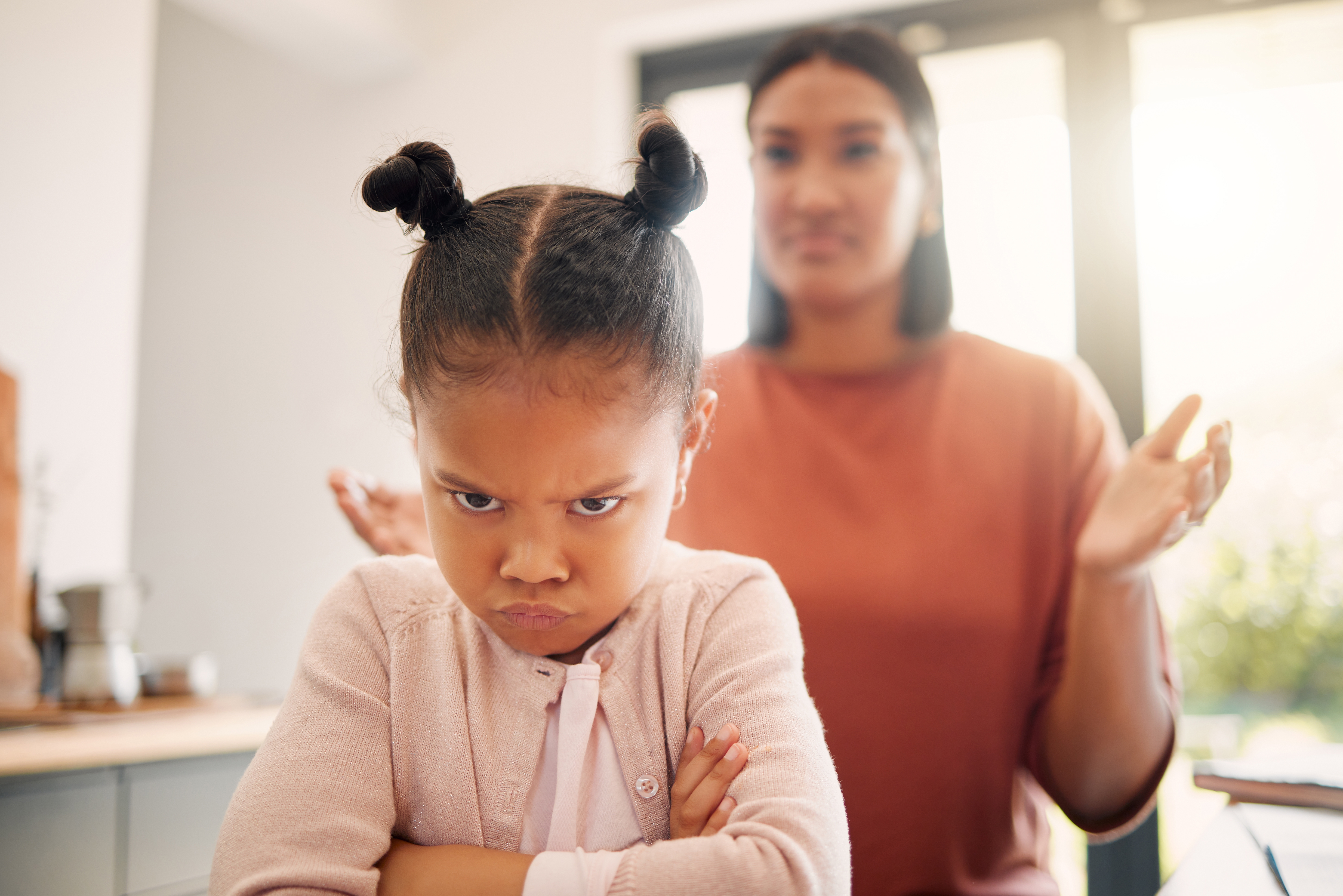 Child with crossed arms and pouty face stands in the foreground, while an adult in the background raises hands in exasperation