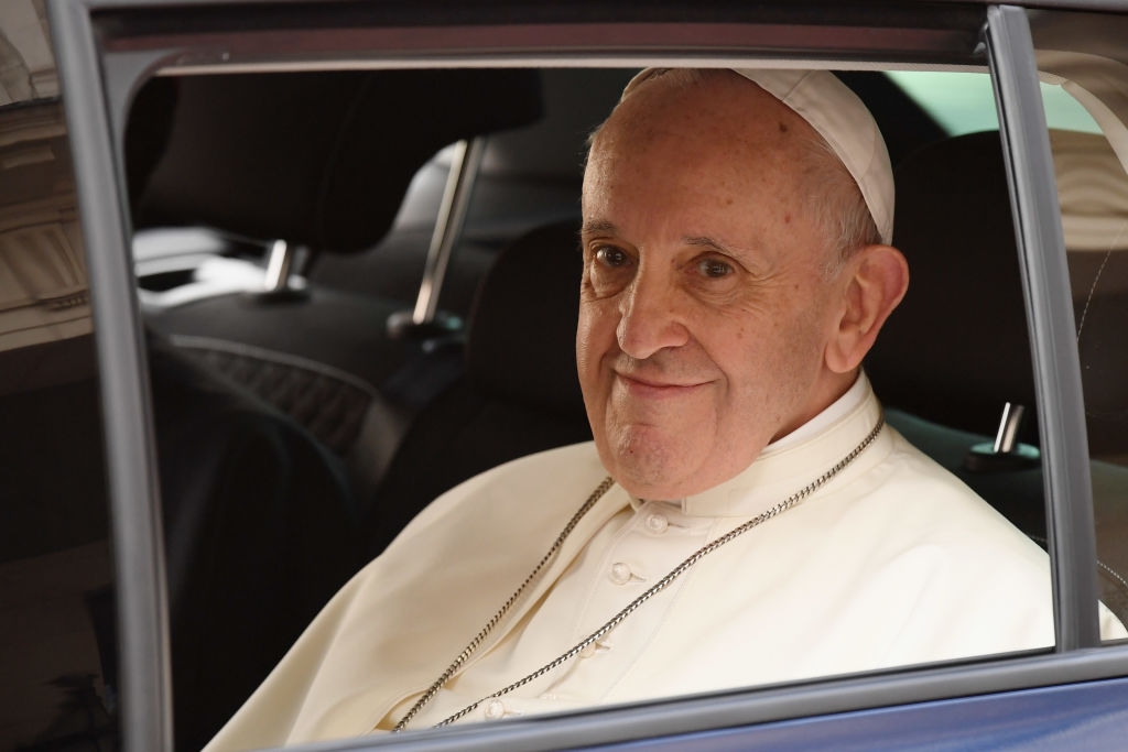 Pope Francis wearing a white religious robe and cap smiles while sitting in a car