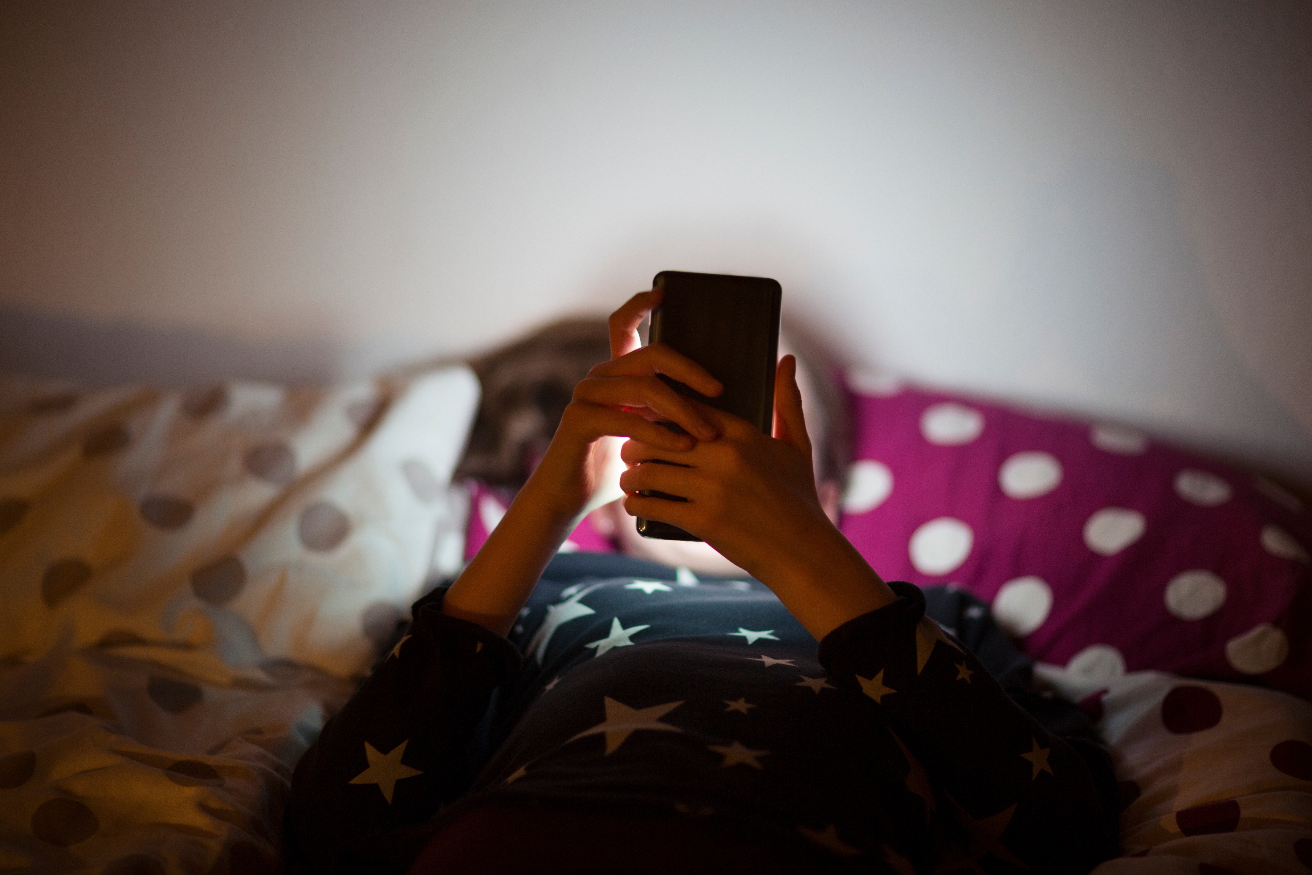 Person in bed looking at a phone. The room is dimly lit, giving a cozy atmosphere. They are covered with a polka dot blanket
