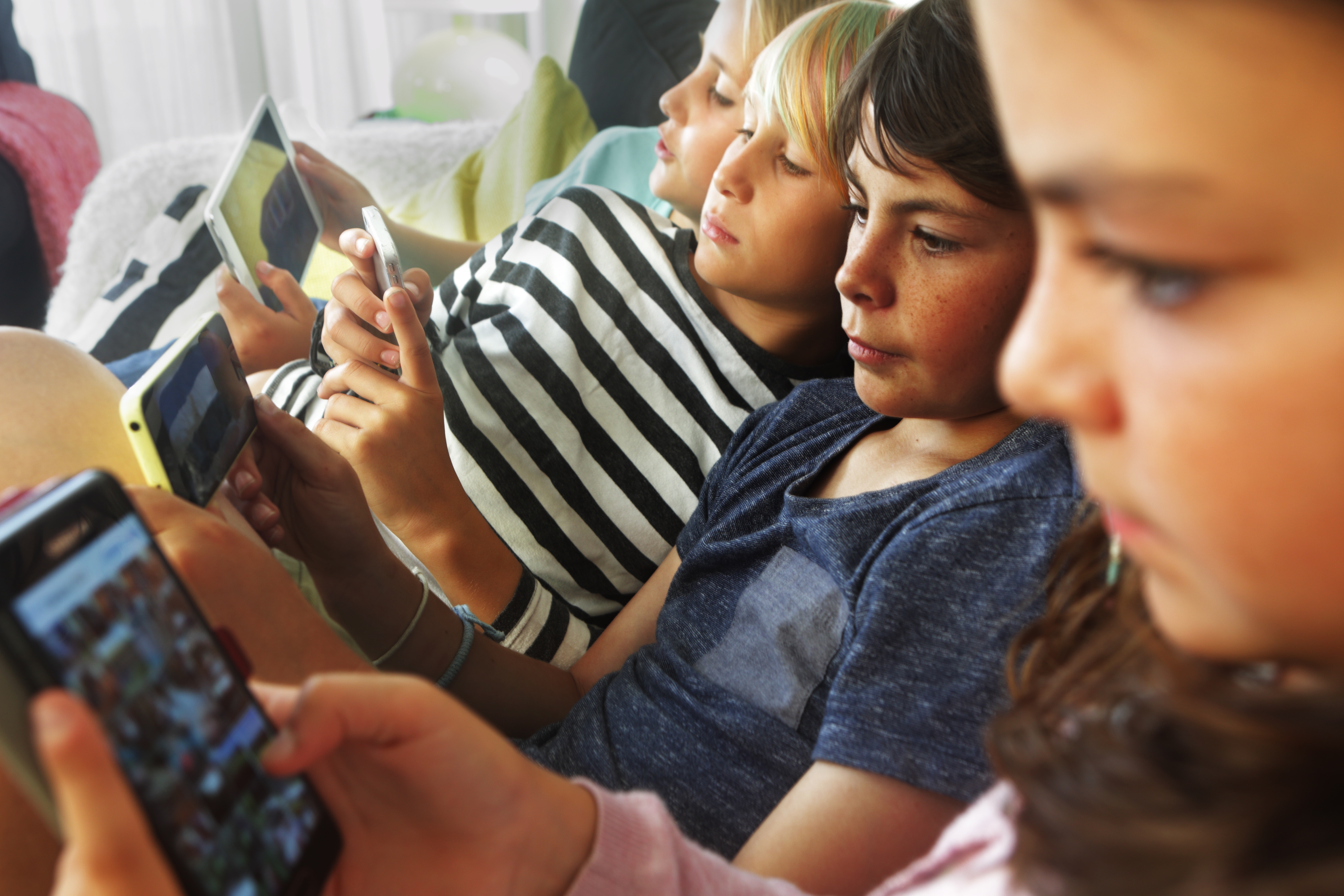 Children lying on a couch, each focused on a smartphone or tablet, engaged in various activities