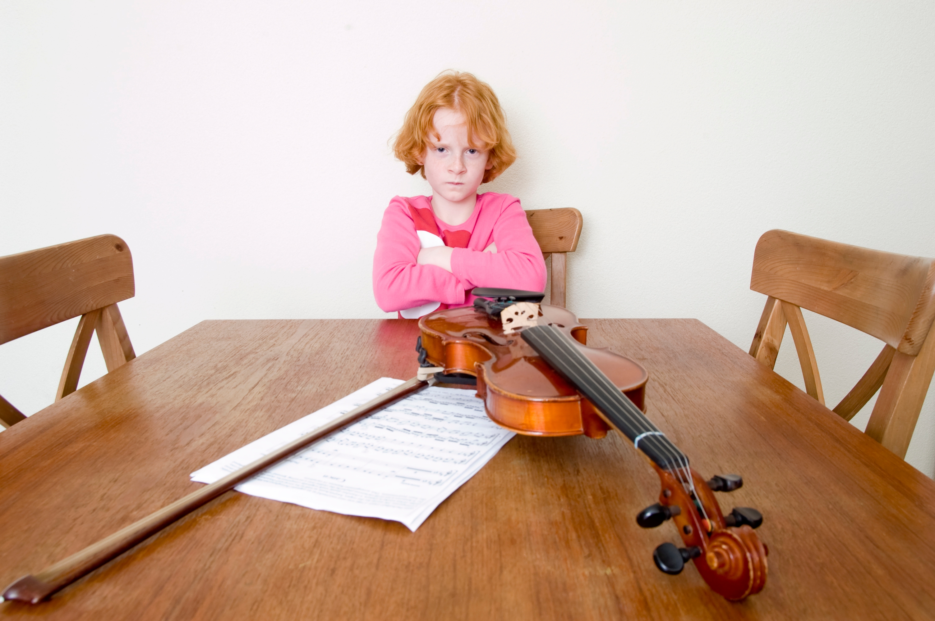 Child with crossed arms sits at a table with a violin and sheet music in front of them