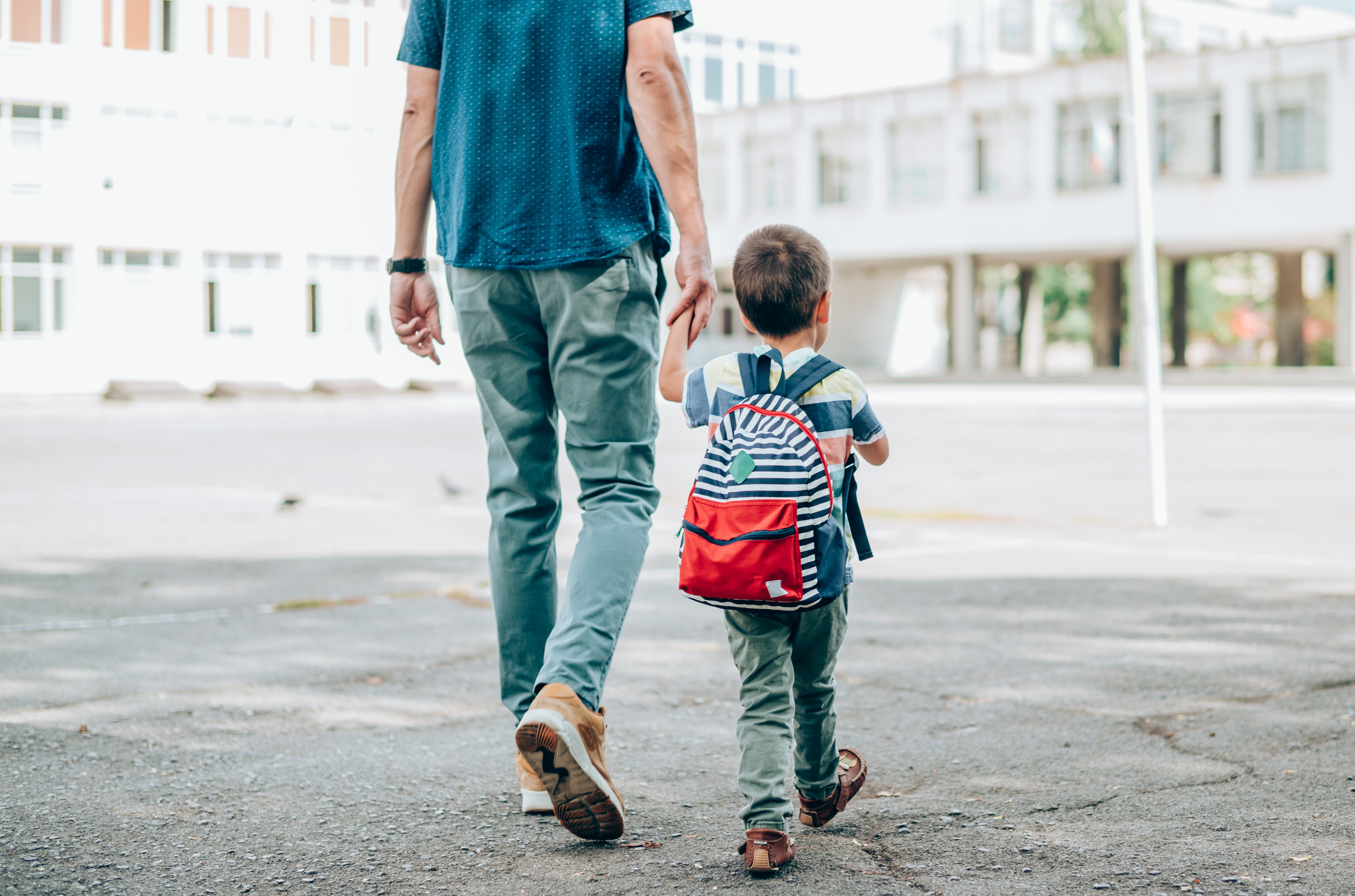 An adult and child holding hands walk on a school campus. The child has a backpack, symbolizing a school day or educational journey