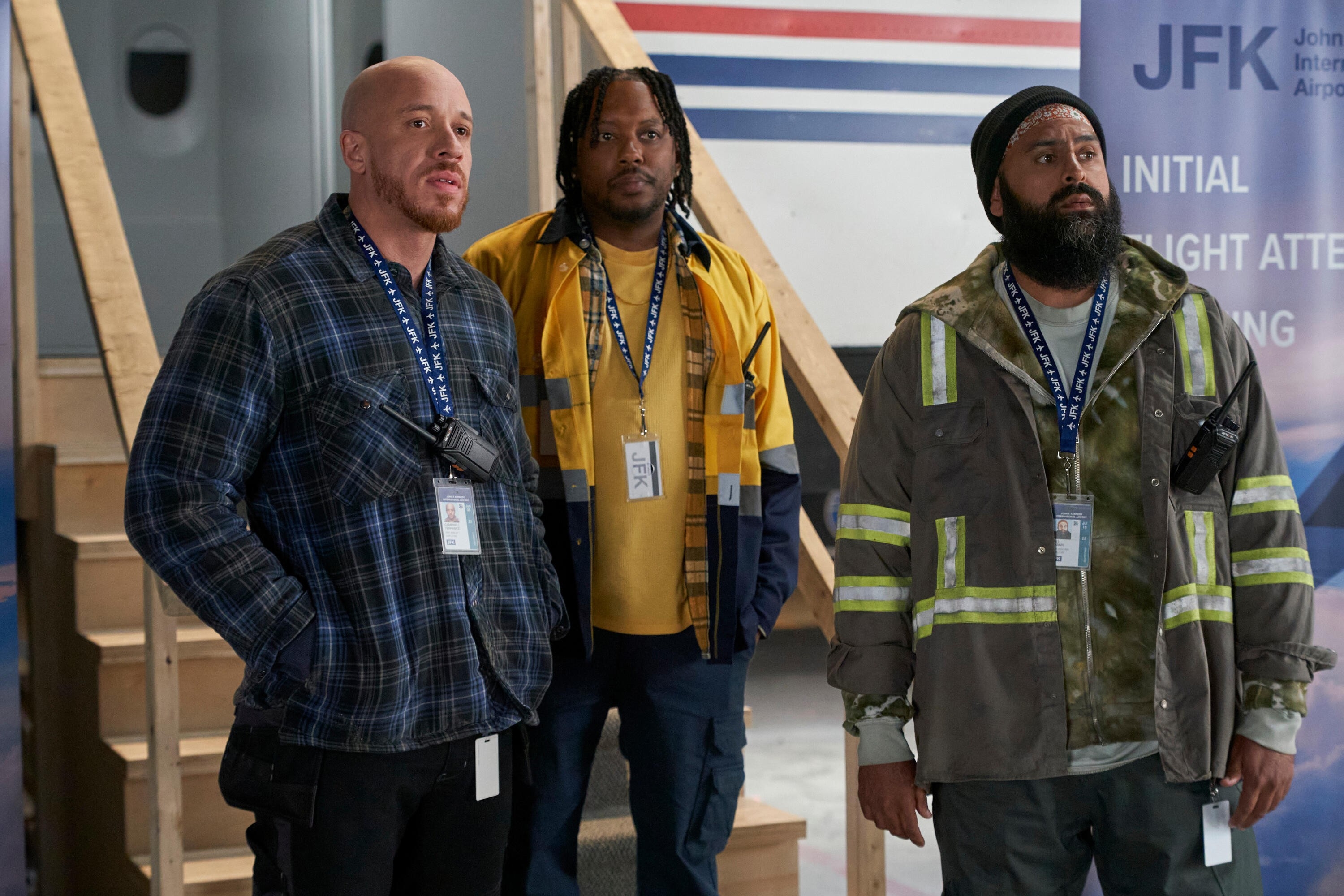 Three actors on a TV set, dressed as airport ground crew, wearing plaid, a yellow jacket, and a reflective work coat, standing in a hangar