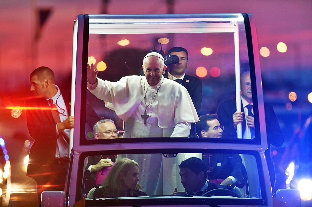 Pope Francis in white robes waves to crowd from a protected vehicle, surrounded by security personnel, in an evening public event
