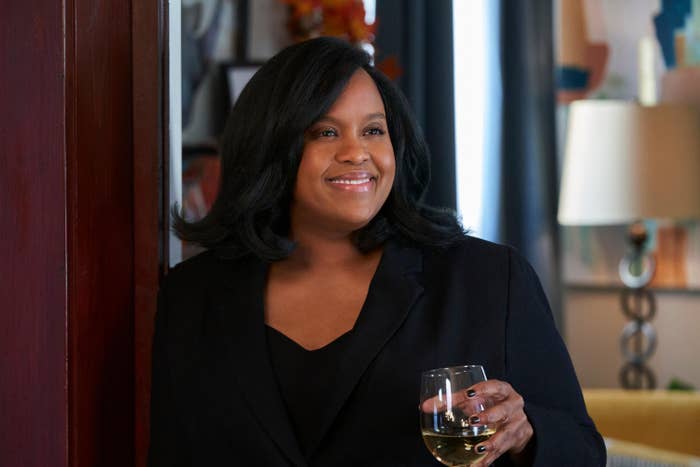 Person smiling in a scene from a TV show, holding a wine glass, wearing a black blazer in a cozy room setting