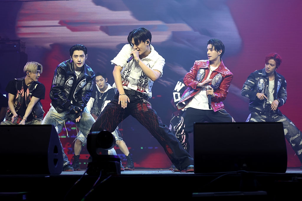 A music group performs on stage, members in dynamic dance poses. They wear stylish, edgy outfits, including jackets and pants, reflecting a modern look