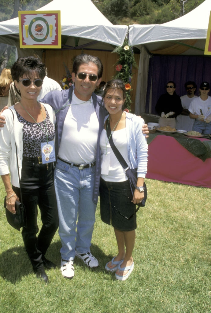 Three people smiling and posing outdoors at an event. The man is wearing a casual jacket and jeans. Tents are visible in the background
