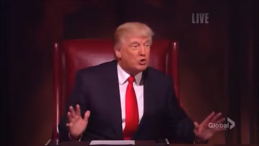A person sits in a leather chair, gesturing with their hands. They're wearing a suit and red tie, appearing to speak on a TV show set