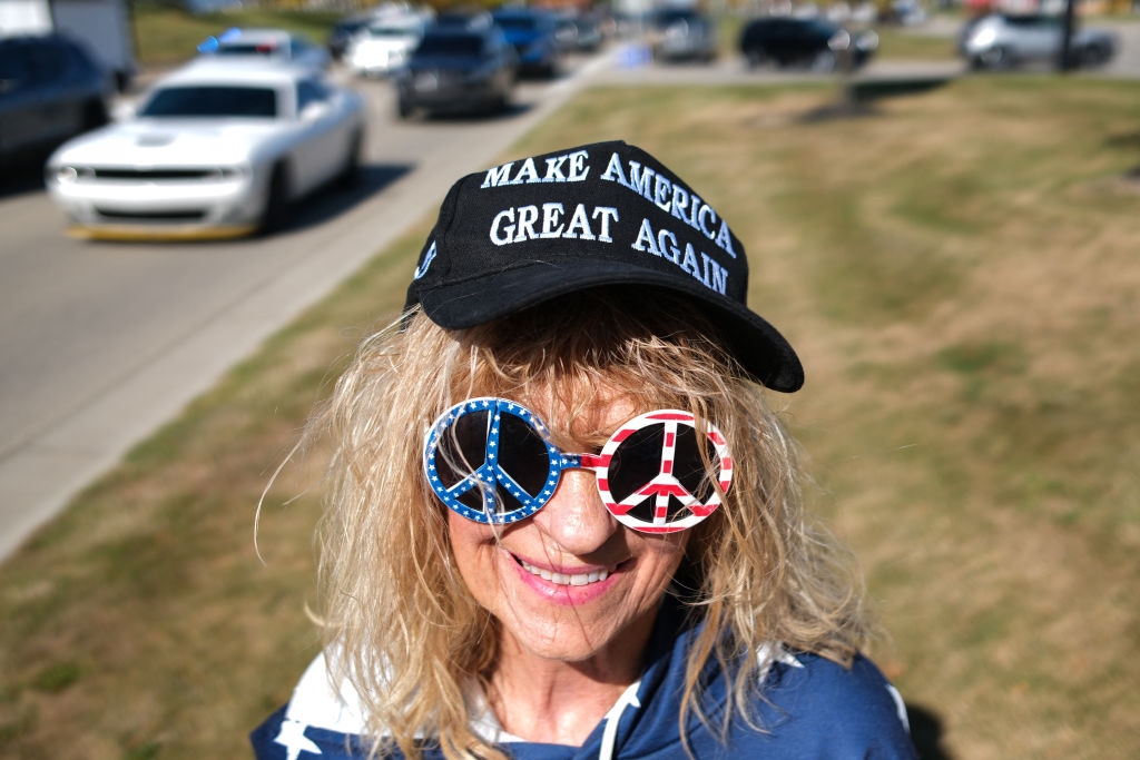 Person wearing peace-sign sunglasses and a &quot;Make America Great Again&quot; hat stands outdoors near a road