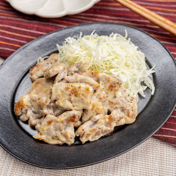 黒い皿に載ったマヨネーズと粒マスタードがからんだ豚こま肉の料理と千切りキャベツ。