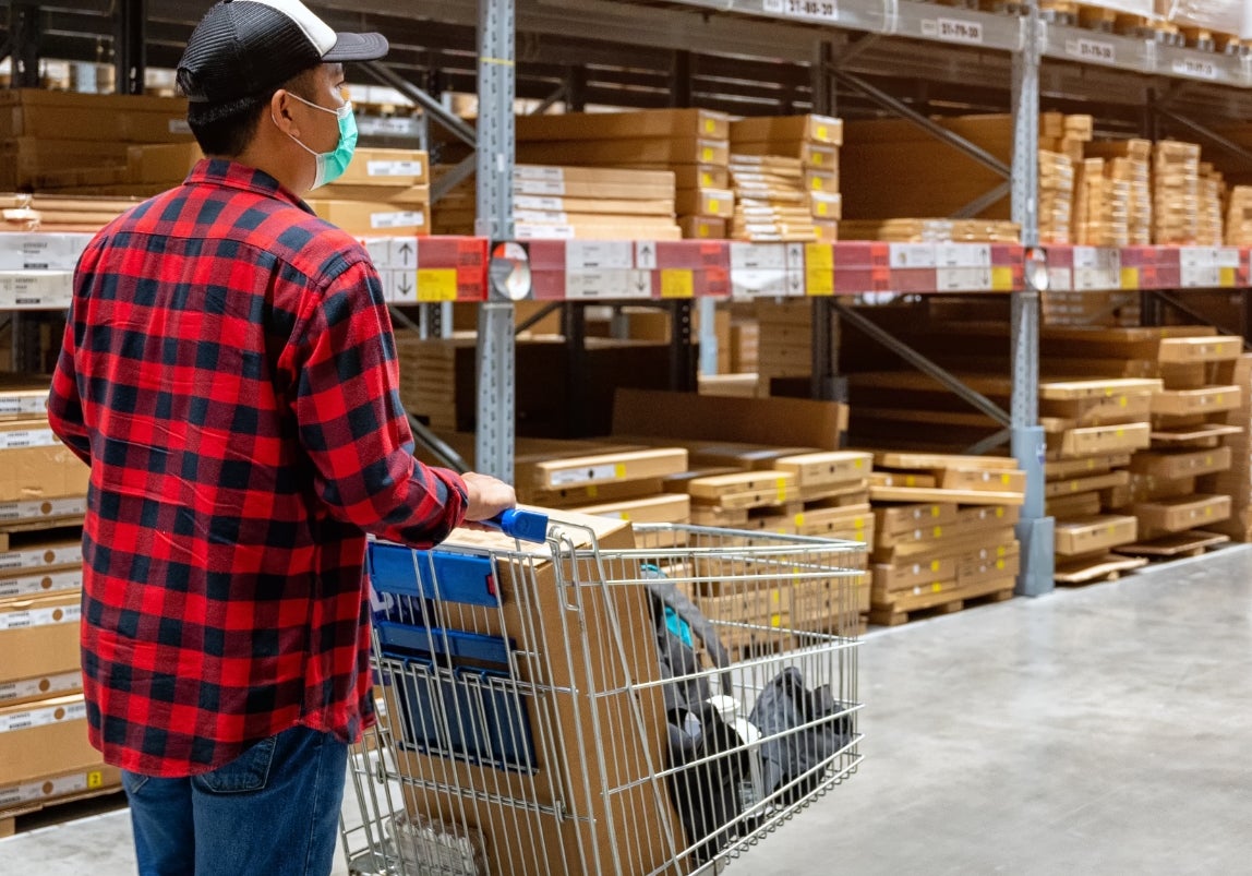 Person in a plaid shirt pushes a shopping cart through a large warehouse filled with boxes and shelves