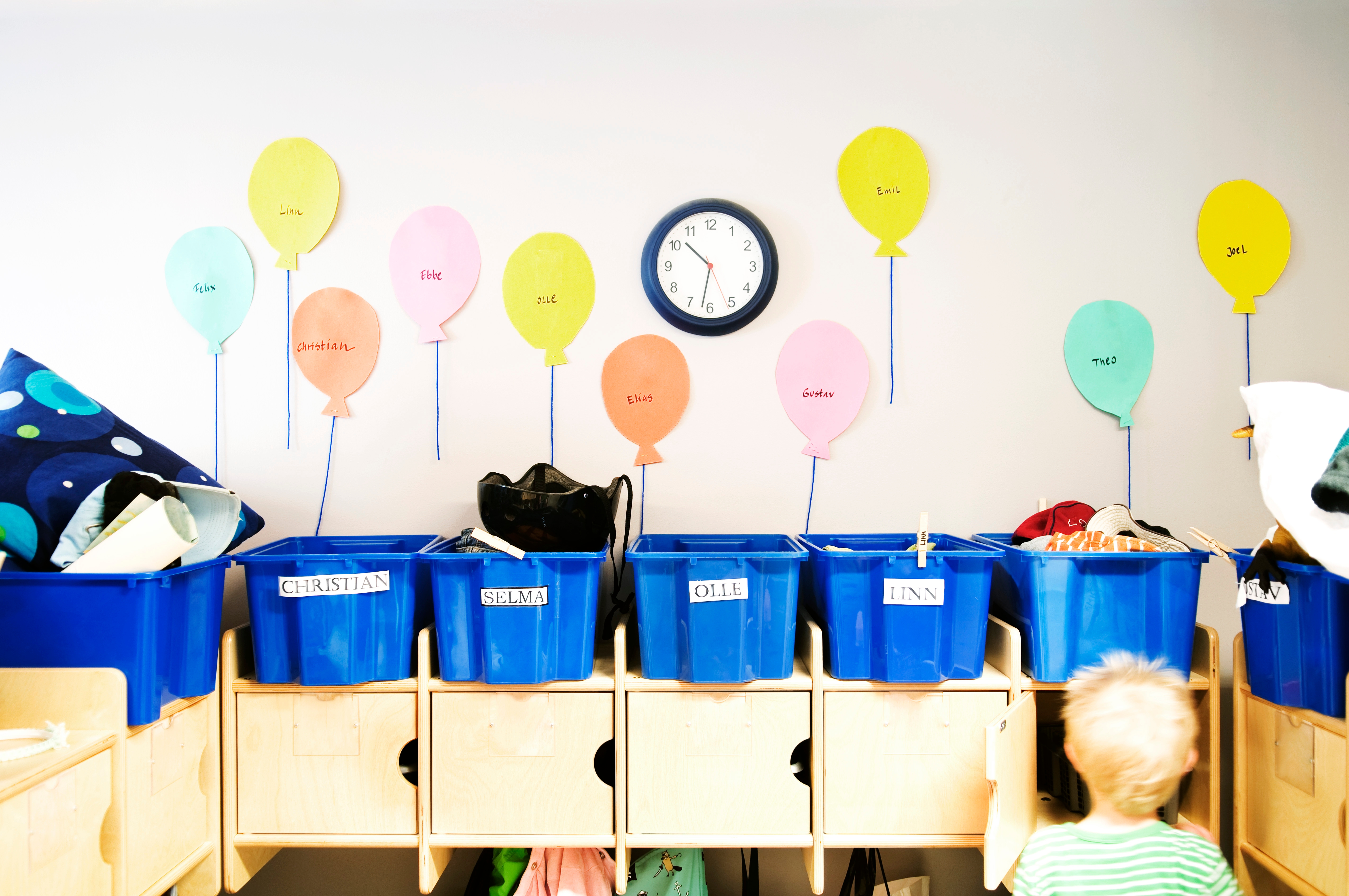 Child in daycare with storage cubbies below. Above, paper balloons on the wall have names. A clock shows 10:10