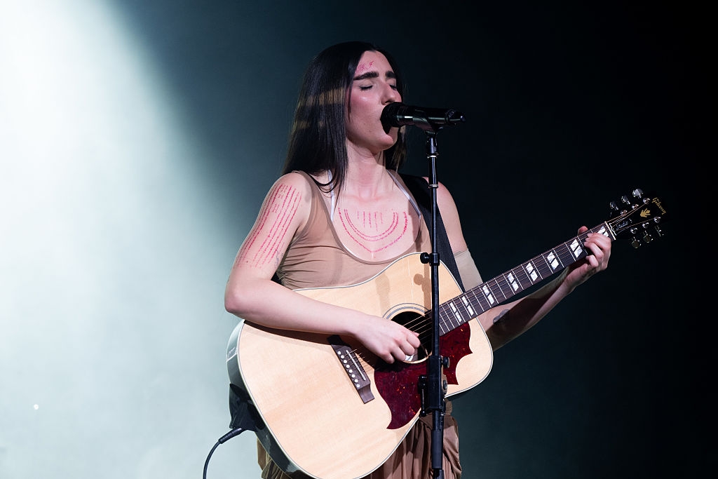 A musician passionately sings while playing an acoustic guitar on stage, wearing a sleeveless dress with geometric designs