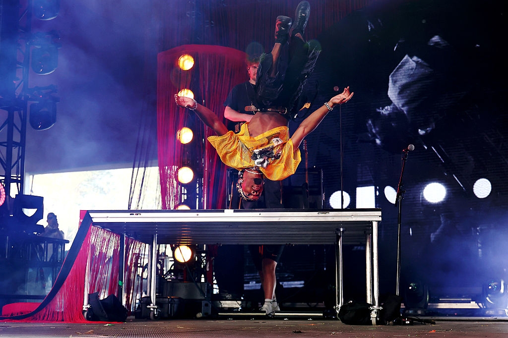 Performer doing a backflip on stage during a live music concert, wearing a graphic t-shirt and jeans. Spectacular stage lights in the background
