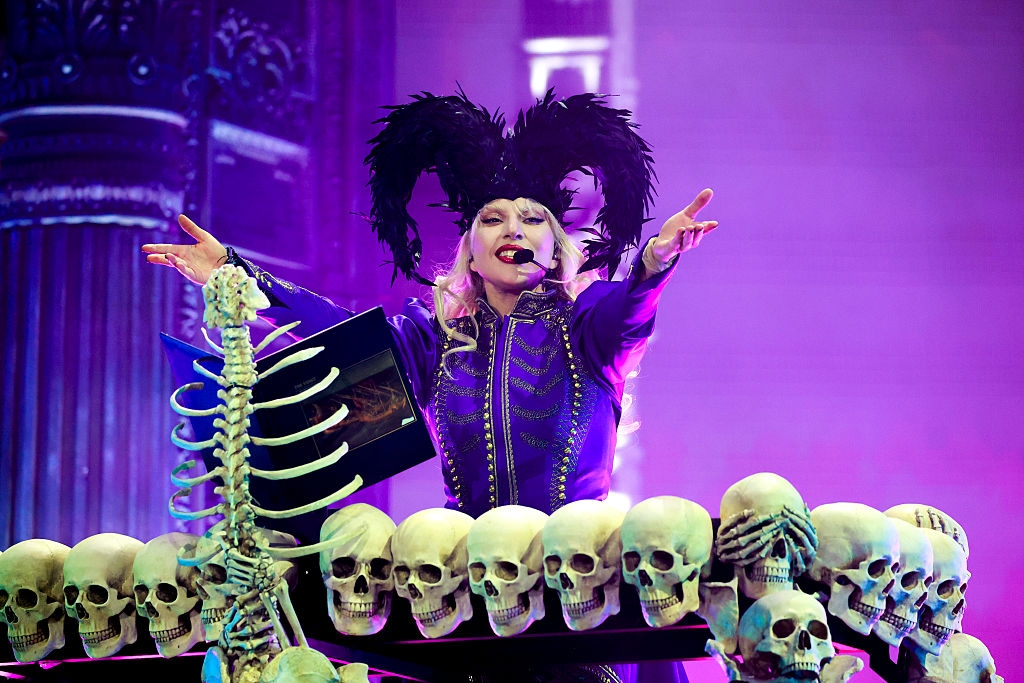 Performer on stage with elaborate feathered headpiece, wearing ornate outfit, surrounded by a decorative setup featuring skulls and skeleton motifs