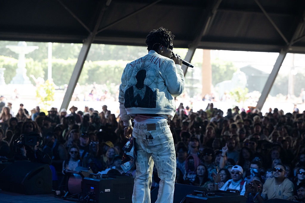 Musician on stage wearing a denim jacket and jeans, performing to a large outdoor crowd at a music festival