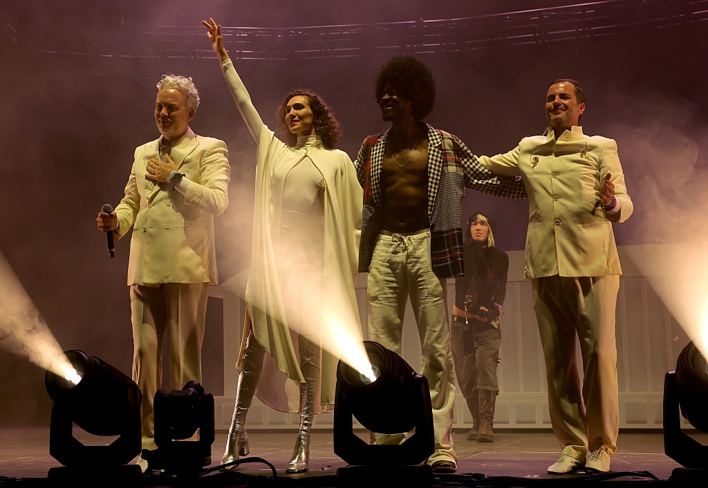 Musicians stand arm-in-arm on stage, dressed in elegant outfits, with one wearing a cape and another shirtless. Stage lights create a dramatic setting