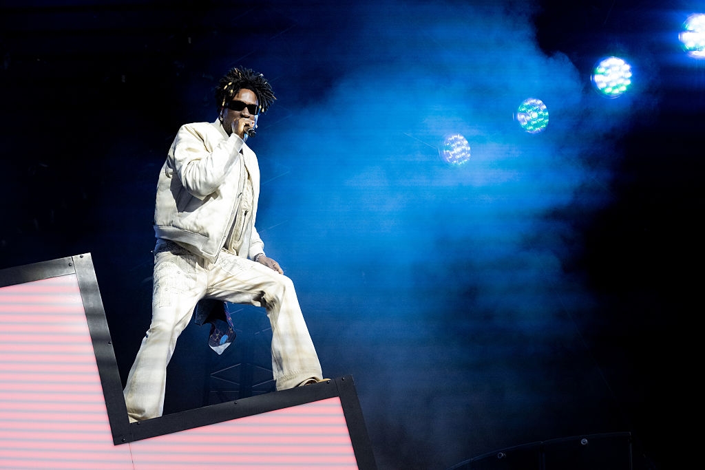 Musician on stage in stylish, oversized outfit and sunglasses, singing energetically amidst dynamic lighting and visuals