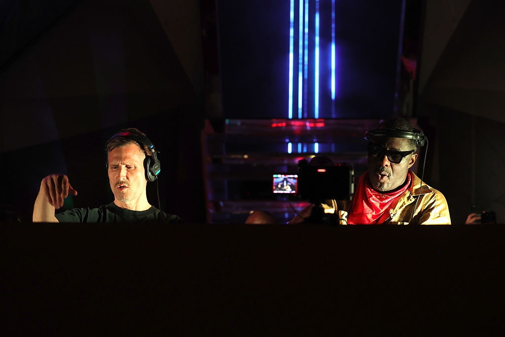 Two DJs performing on stage, wearing headphones, in front of electronic equipment, under dim lighting