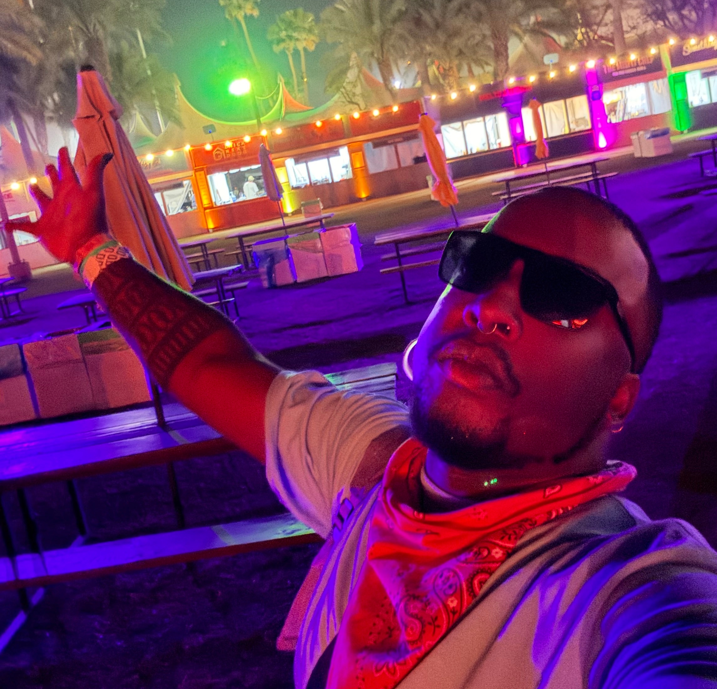 Person wearing sunglasses and a bandana poses energetically at an outdoor music festival during the night, with lights and palm trees in the background