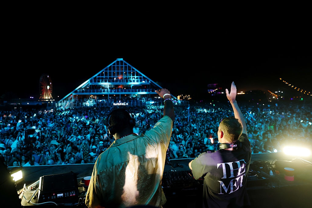 Two DJs perform on an outdoor stage at a music festival, facing a large, energized crowd. One DJ wears a jacket, the other wears a printed T-shirt