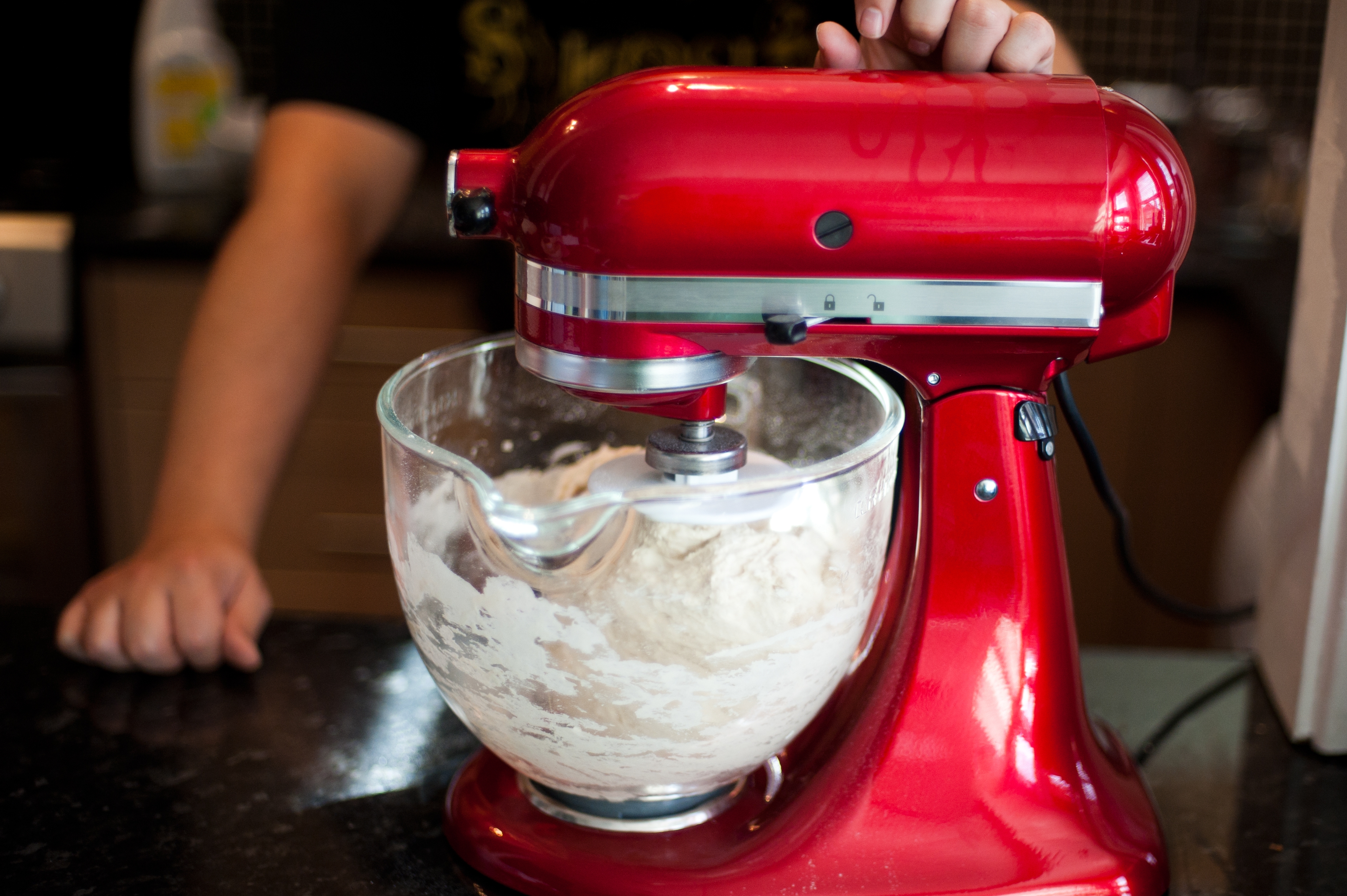 A person operates a red stand mixer with dough in a glass bowl on a kitchen counter