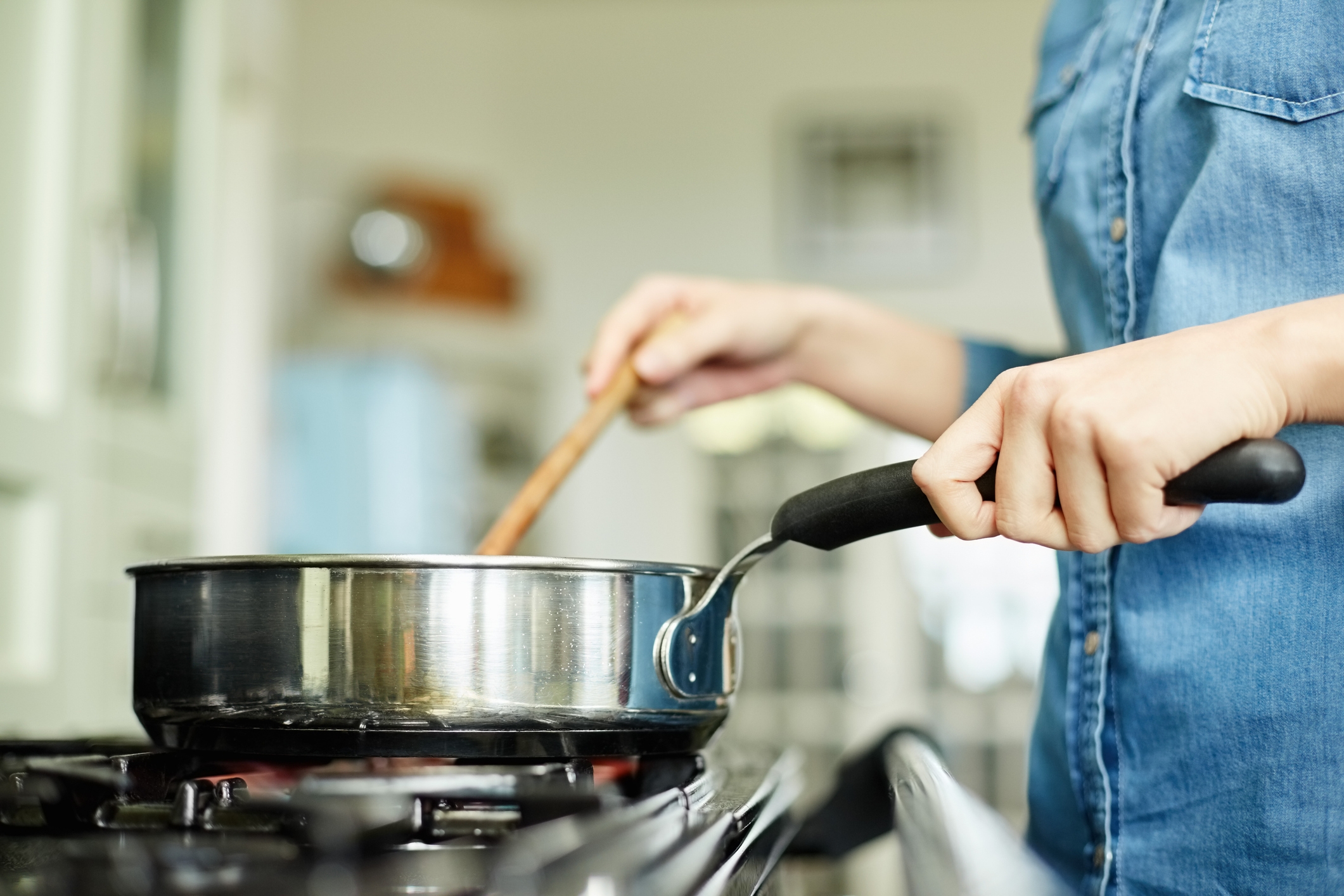 Person stirring pot on a stove with a wooden spoon, wearing a casual denim shirt. Cooking in a home kitchen setting