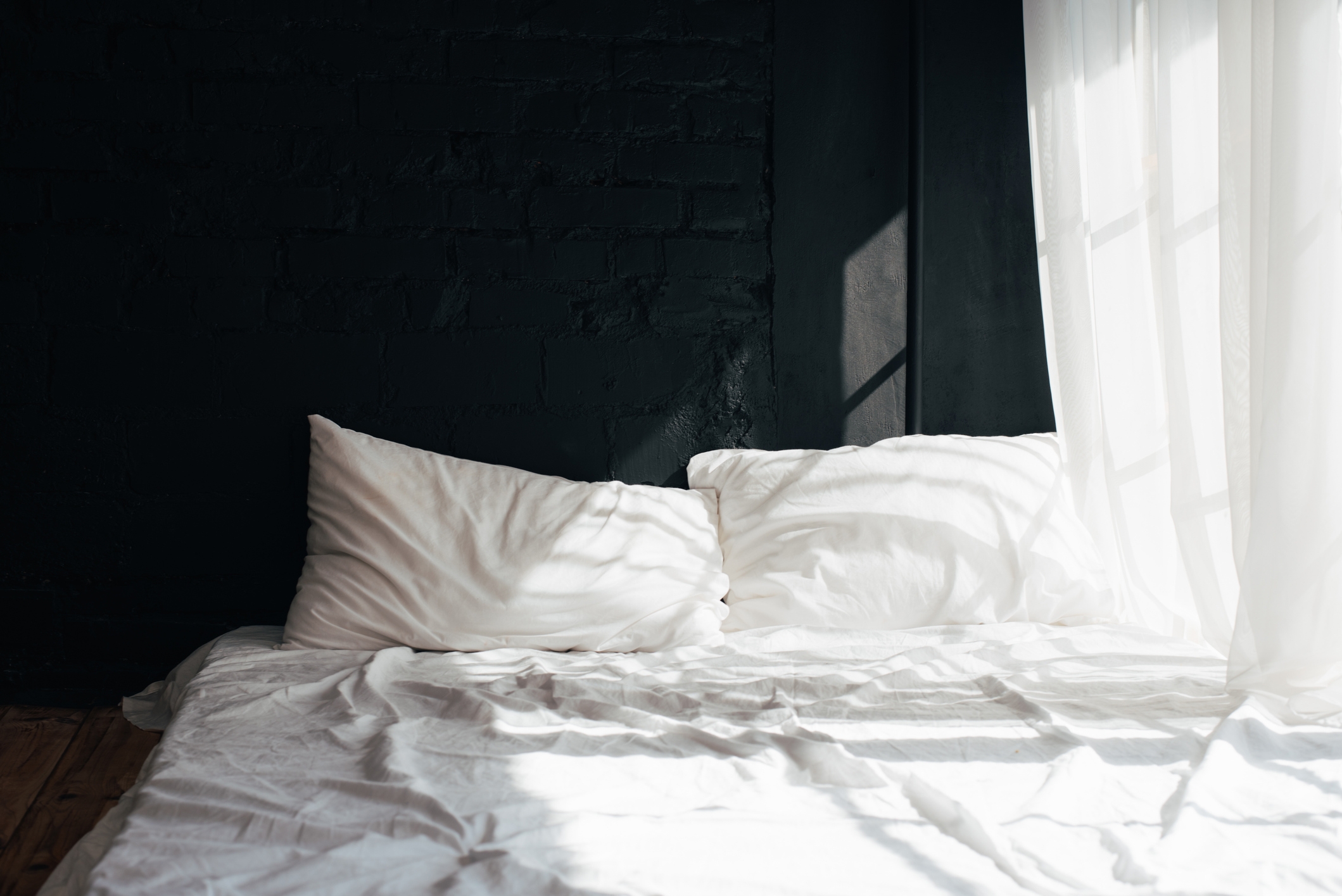 A neatly made bed with white pillows and sheets next to sheer curtains, letting in soft, filtered light from a window