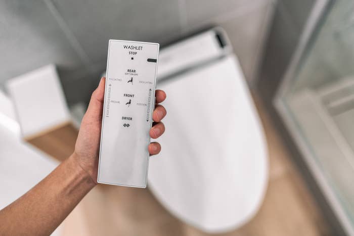 A hand holds a remote control for a bidet toilet, showing options for rear, front washing, and drying