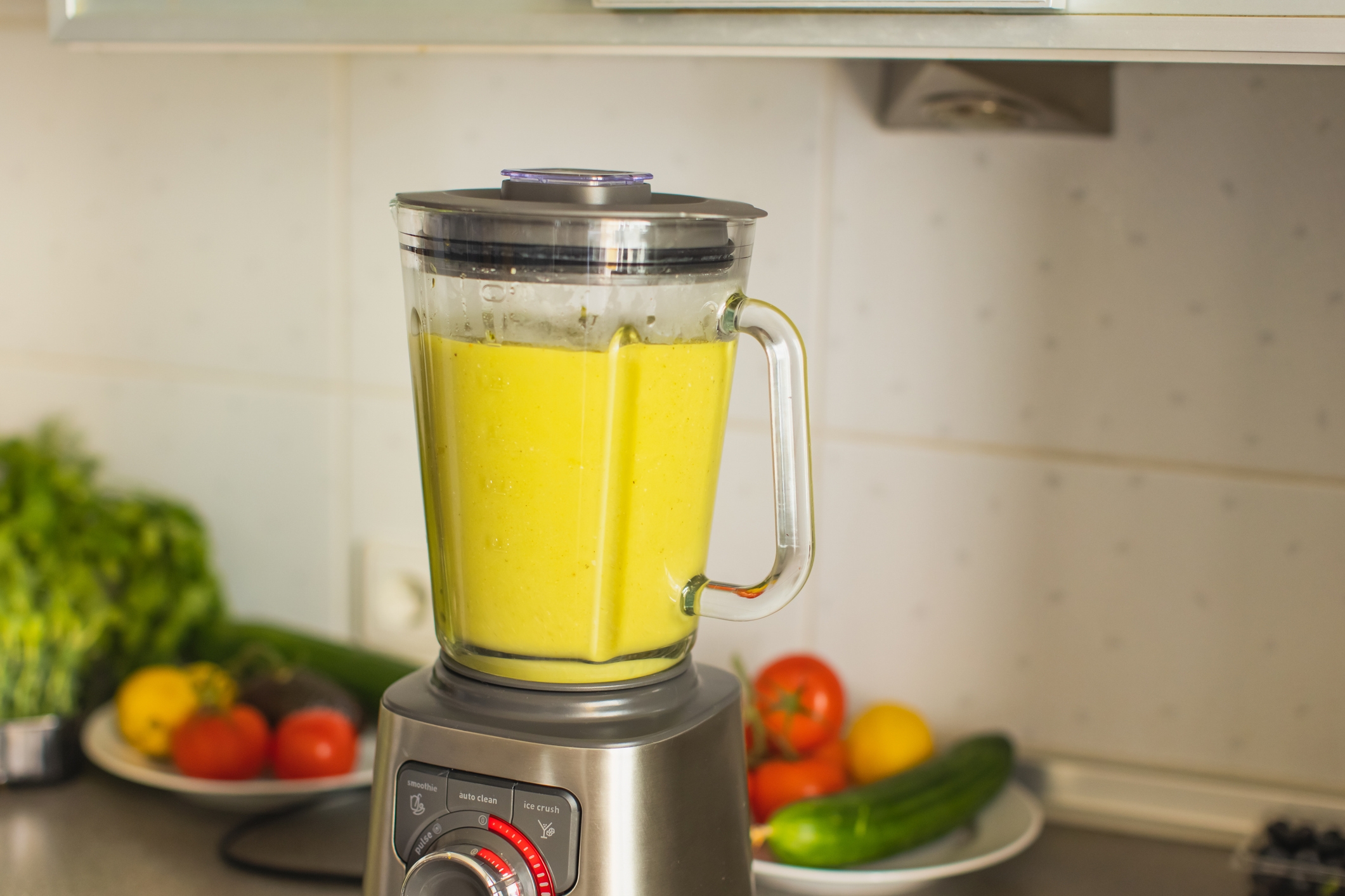 Blender on kitchen counter with yellow smoothie inside, surrounded by assorted fresh vegetables and fruits on plates in the background