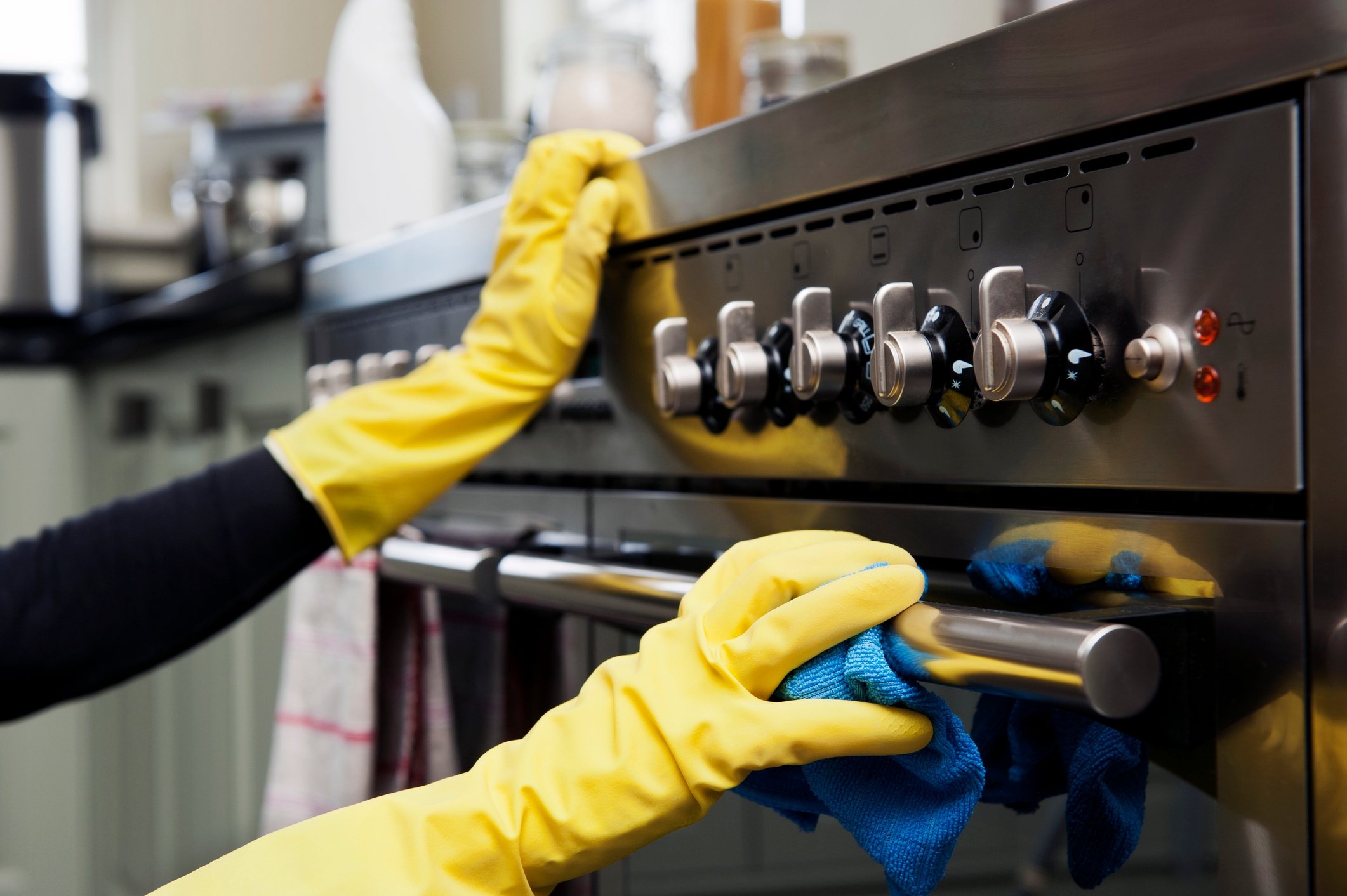 Person wearing yellow gloves cleaning an oven with a blue cloth, focusing on knobs and a handle
