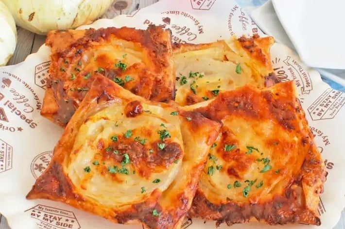 パイ生地に玉ねぎとチーズが乗った焼き料理が4つ皿に盛られている。背景にタマネギと花が飾られている。