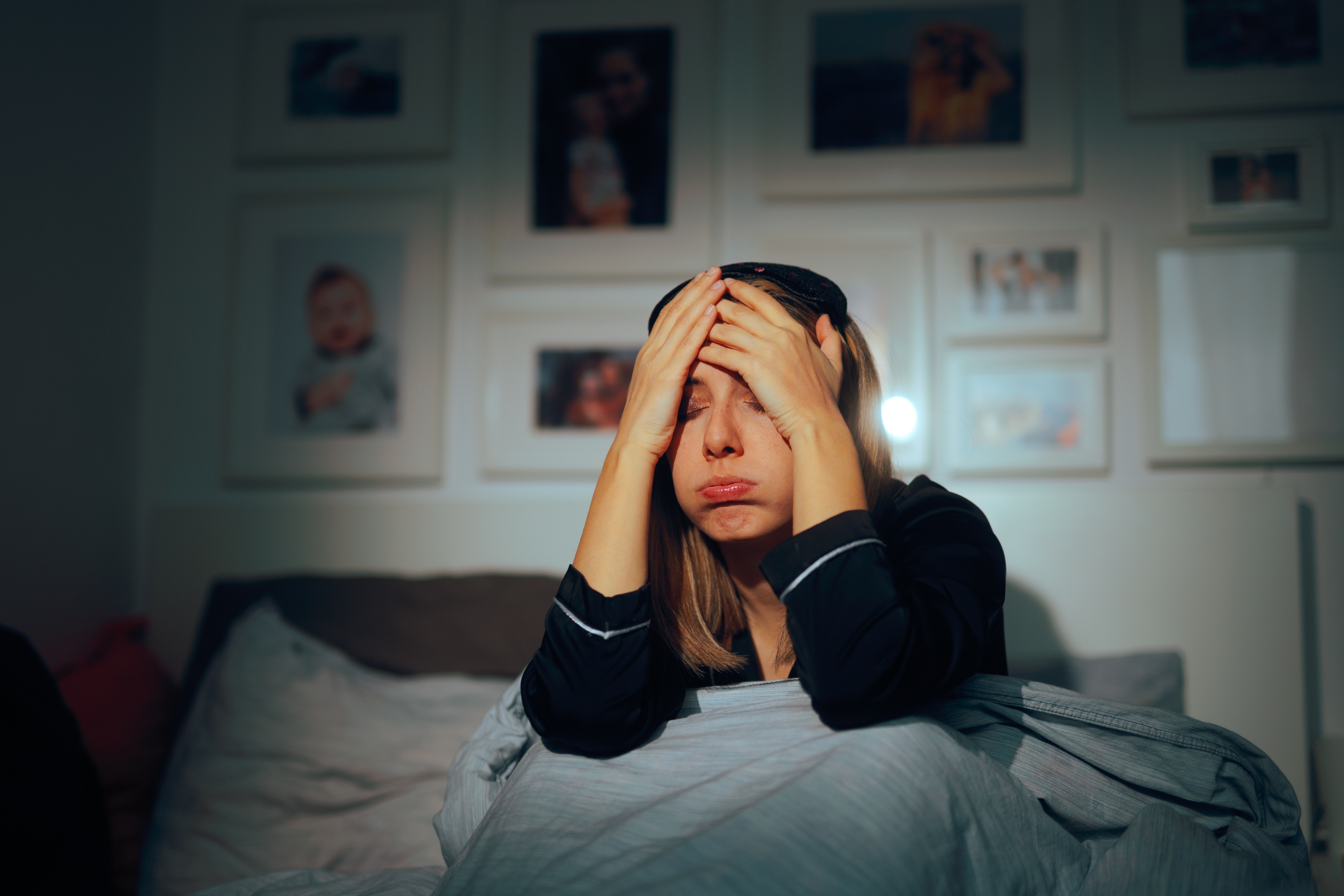 Person sitting on a bed, head in hands, appearing tired or stressed, in a room with framed photos on the wall