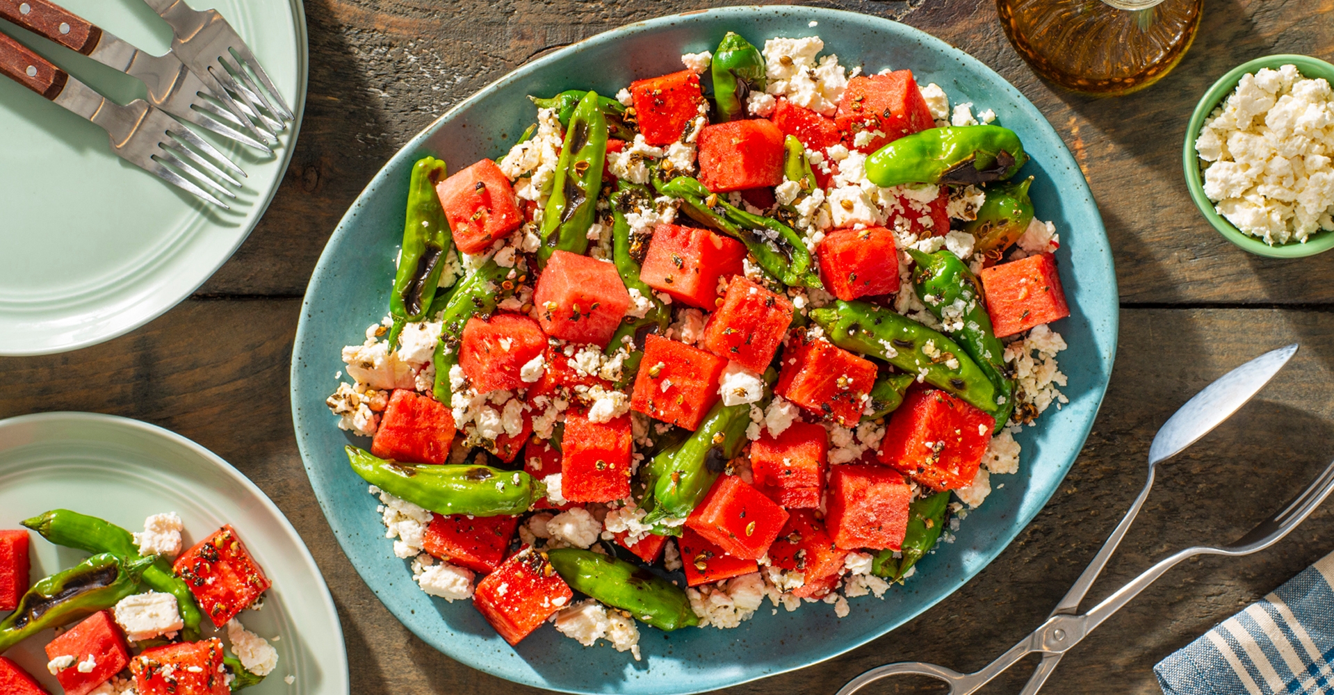 A platter of watermelon, feta cheese, and green peppers, garnished with herbs, on a table set with plates and cutlery