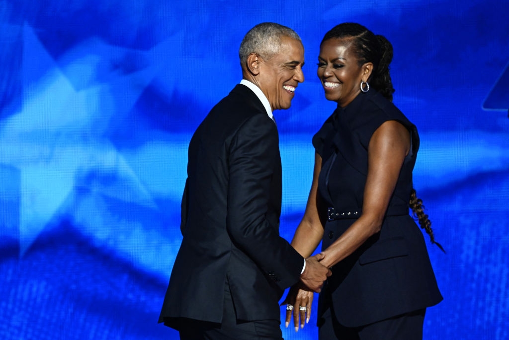 A couple smiling warmly, holding hands on stage, both dressed in elegant black formal attire