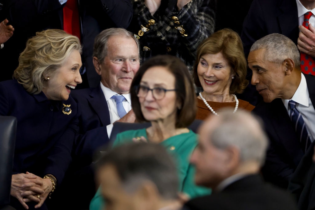 Several public figures converse and smile together at a formal event, wearing suits and elegant attire. Audience members are seated around them