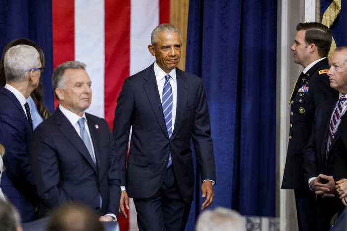 A public figure in a suit walks past others in suits and military uniforms, with an American flag backdrop, at a formal event