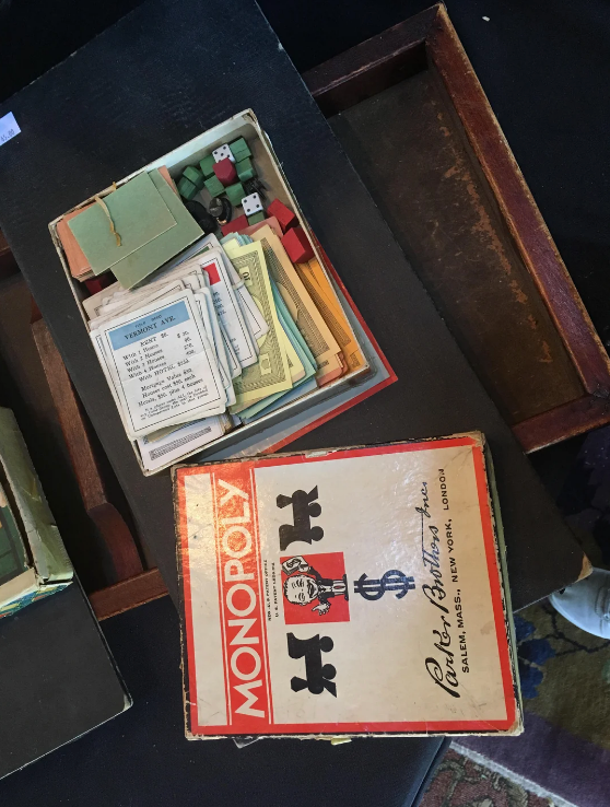 Open vintage Monopoly game with scattered pieces, cards, and dice. Box reads "Monopoly" with London manufacturer's name visible and an old man holding money