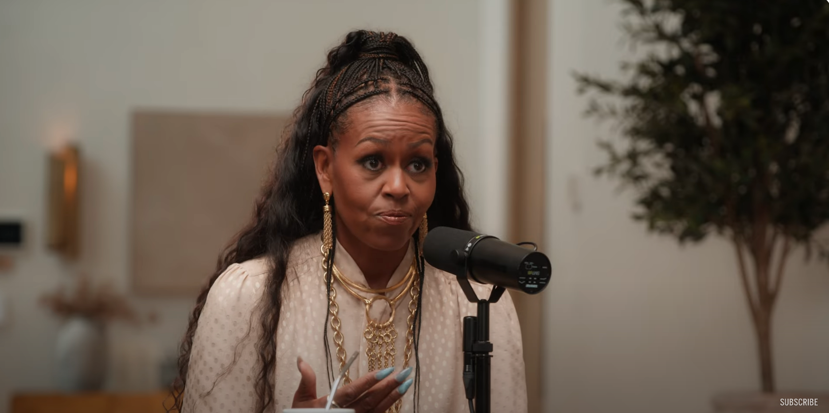 A person with long hair is speaking into a microphone, wearing a white outfit with layered necklaces