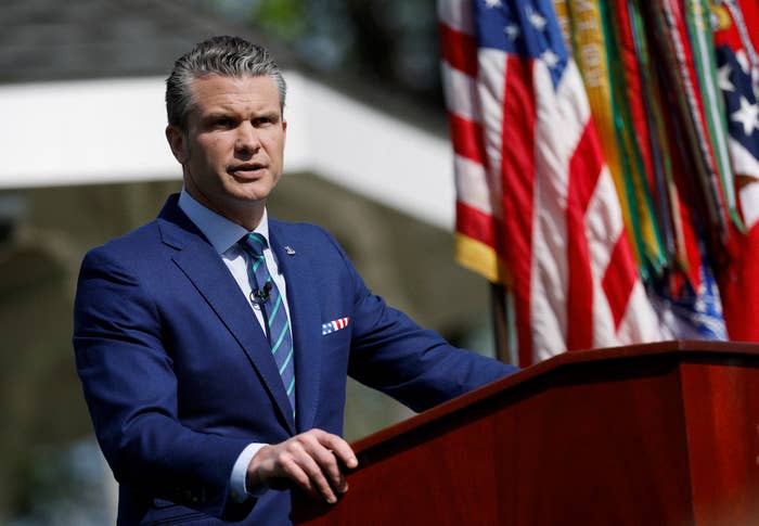 A person in a suit stands at a podium outdoors, speaking. Flags are in the background