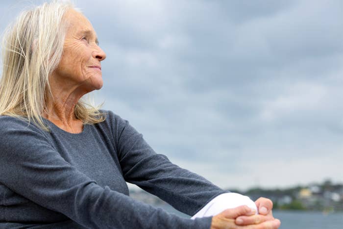 Older woman with long hair sits outdoors, relaxed and contemplative, against a cloudy sky