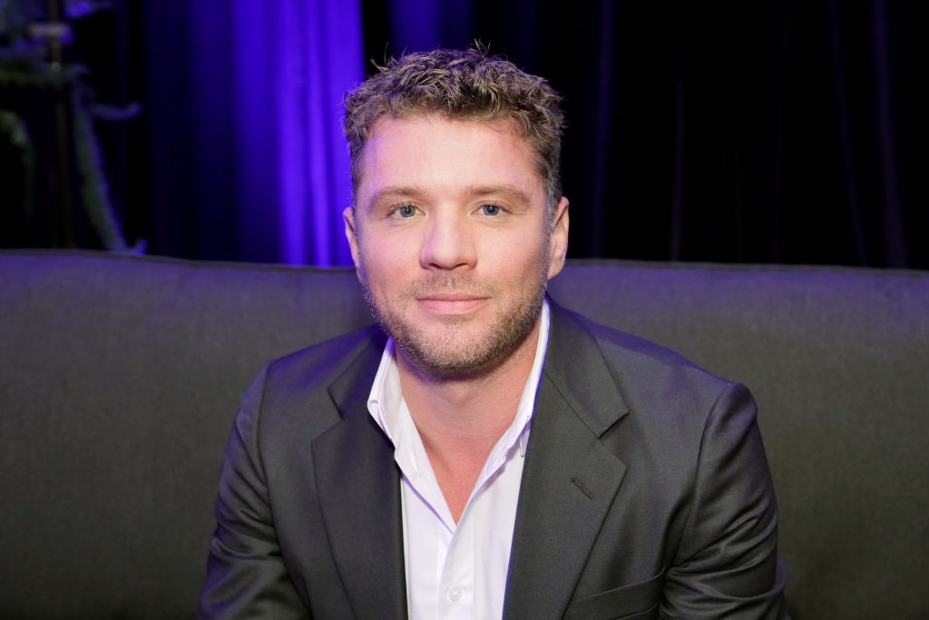 A man in a black suit and white shirt sits on a dark couch, smiling slightly, with a purple-lit background