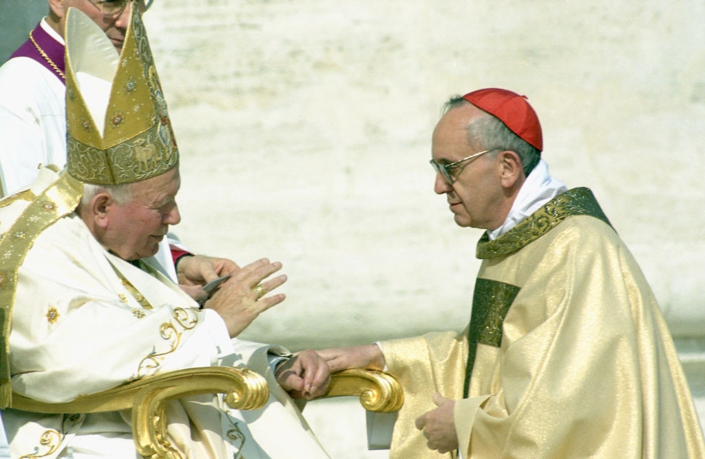 Cardinal Jorge Bergoglio kneels before Pope John Paul II in formal attire engaged in conversation, seated, with one wearing a mitre and the other a biretta