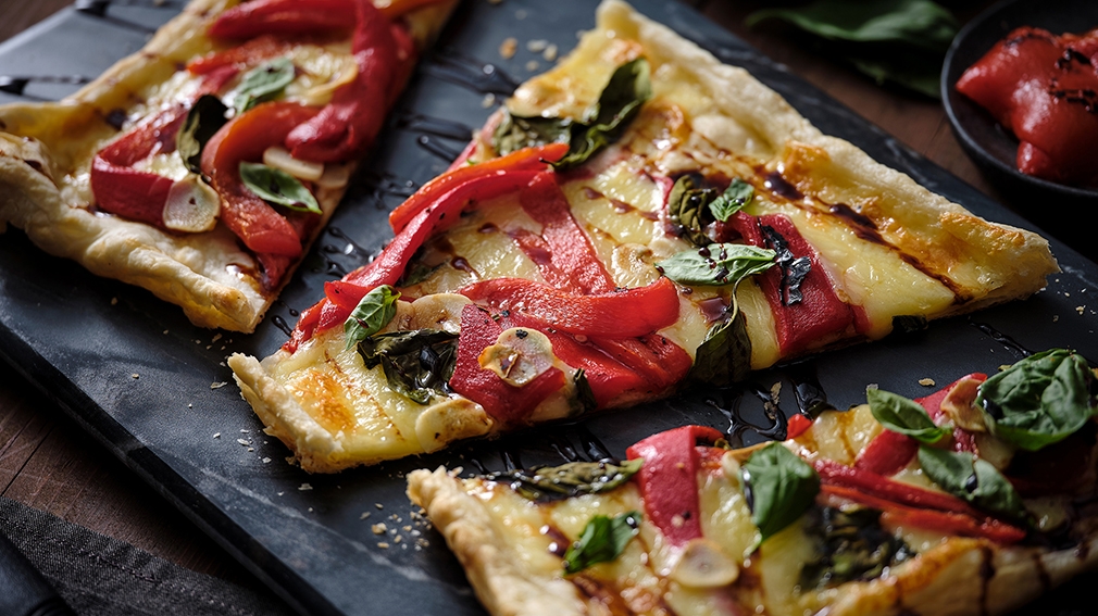 Slices of pizza topped with red peppers, basil, and a drizzle of balsamic glaze on a slate board