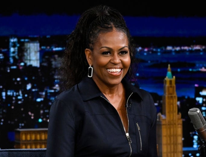 Person on a talk show set, wearing a chic black outfit with hoop earrings, sitting and smiling