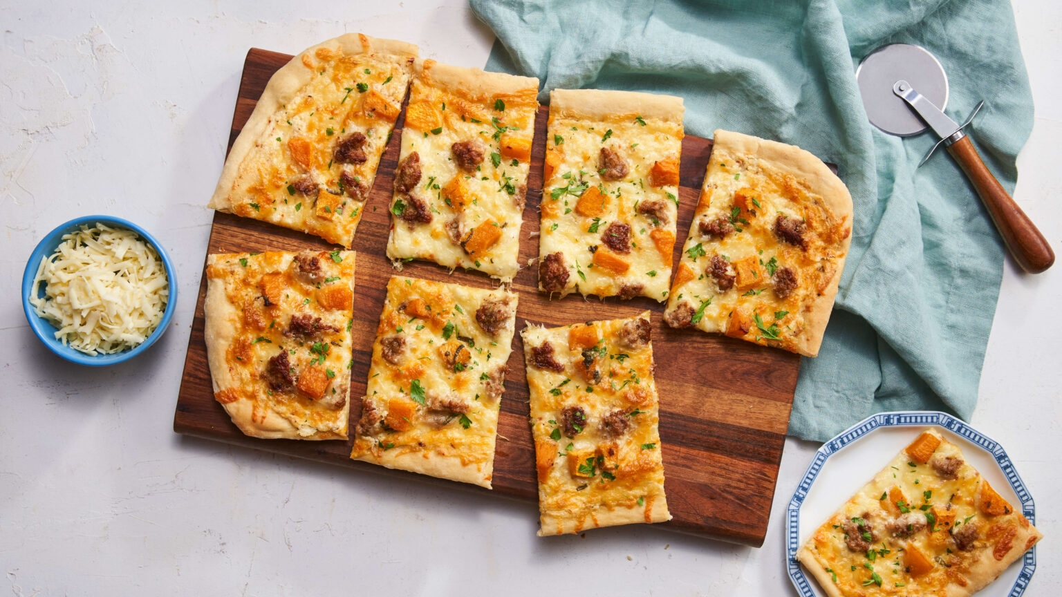Flatbread pizza with sausage, cheese, and vegetables cut into rectangles on a wooden board, next to a bowl of grated cheese and a pizza cutter