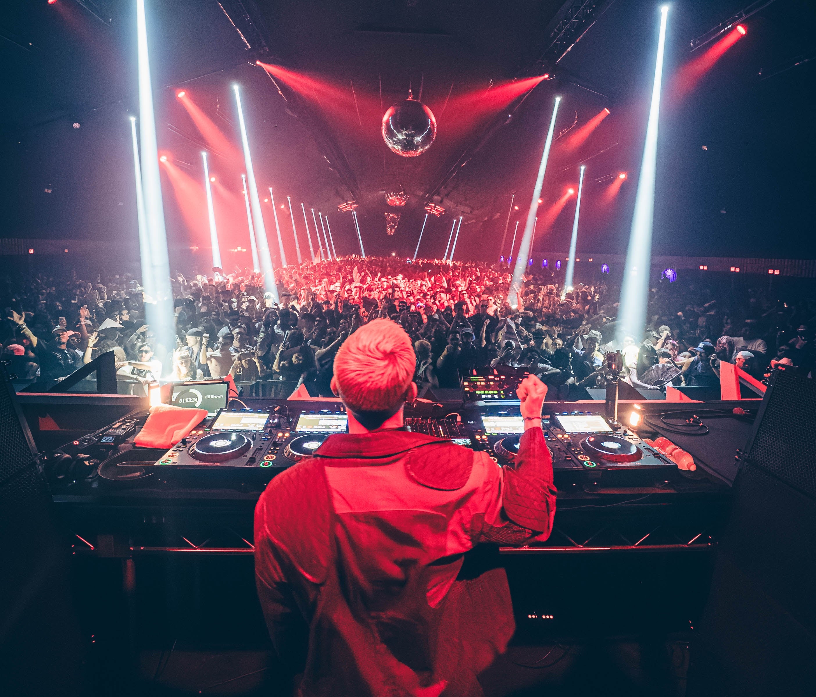 DJ performing live in front of a large, energetic nightclub crowd with vibrant stage lights and a disco ball overhead