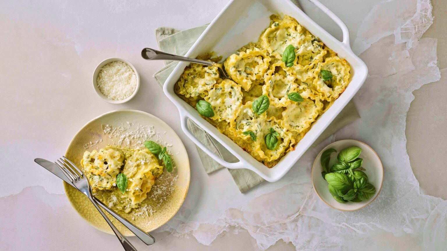 A dish of lasagna with basil garnish in a baking dish, accompanied by a plated serving, grated cheese, and fresh basil in small bowls
