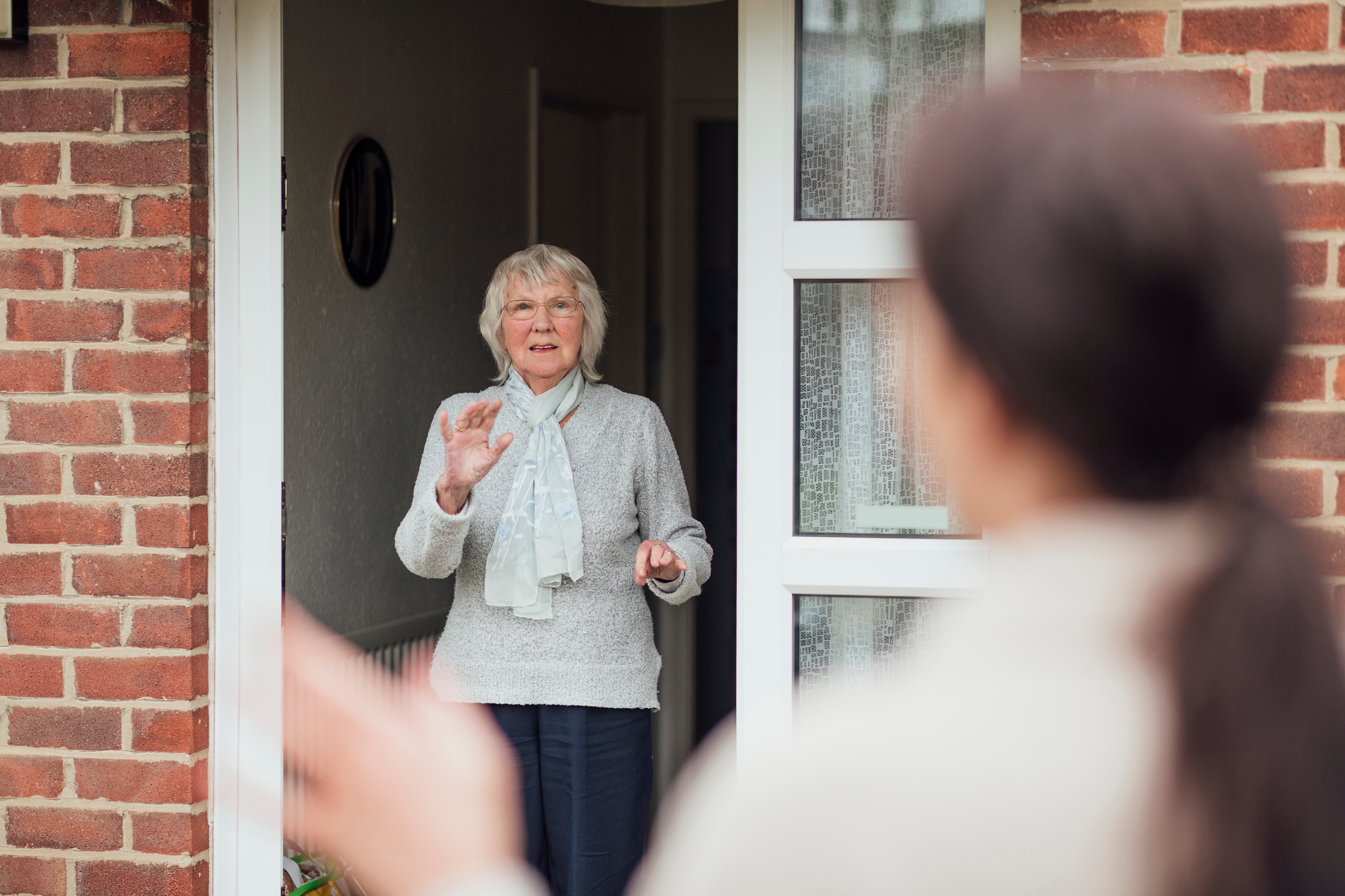 An elderly woman stands at her home's open door, smiling and waving at a visitor with blurred hands in the foreground
